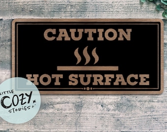 May include: A rectangular wooden sign with a black background and the words "CAUTION HOT SURFACE" in a light brown, rustic font. The sign includes a graphic of three steam lines above a horizontal line.