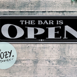 Op de afbeelding: Een rechthoekig zwart en zilveren bord met de woorden "THE BAR IS OPEN" in grote, vette letters. Een kleine, ronde sticker met de tekst "LITTLE COZY STORIES+" staat in de linker benedenhoek. Het bord is bevestigd aan een verweerde, grijze houten muur.