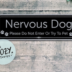 Può includere: Un cartello rettangolare nero con testo bianco che recita Nervous Dog Please Do Not Enter Or Try To Pet. Il cartello presenta grafiche di impronte di zampe bianche sui lati sinistro e destro e un elemento decorativo a linea centrato.