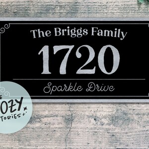May include: A rectangular black and silver address sign with the text "The Briggs Family", the number "1720", and "Sparkle Drive". The sign has decorative corner accents and a silver border. The sign is mounted on a textured grey wall.