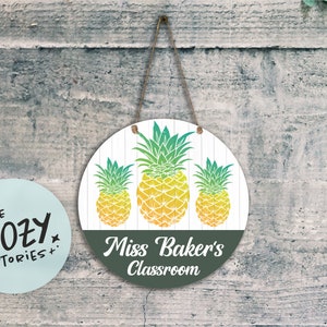 Op de afbeelding: Een rond houten bord met een groen, geel en wit ananasontwerp. Het bord heeft een donkergroene achtergrond met de tekst "Miss Baker's Classroom" in wit.
