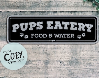 May include: A black and silver sign with the text "Pups Eatery Food & Water" and two paw prints on either side of the text. The sign is mounted on a wooden wall.