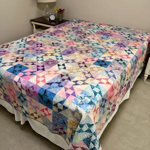 May include: A patchwork quilt featuring a repeating geometric star pattern in shades of pink, purple, blue, and white. The fabric displays a variety of mottled and textured prints, creating a multi-colored design that covers the entire bed surface.