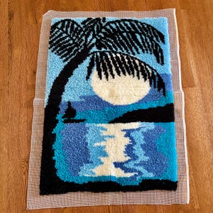 May include: A rectangular latch hook rug featuring a tropical scene with a large black palm tree silhouette against a light blue sky and a white full moon. The design includes a dark blue ocean, a small black sailboat, and a reflection of the moon.