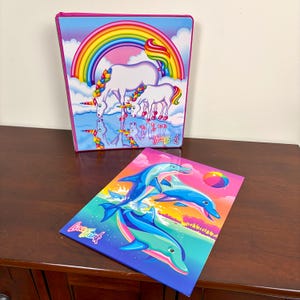 May include: A Lisa Frank three-ring binder featuring a white unicorn and foal under a rainbow, and a matching folder with blue dolphins jumping over water. Both items display the Lisa Frank logo in colorful script against vibrant backgrounds.