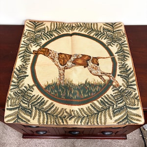 May include: A square decorative pillow featuring a hunting dog design. The dog is brown and white, set against a cream background within a green circle. The border is adorned with green and brown fern-like patterns.