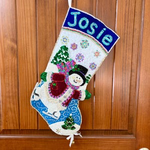 May include: A white Christmas stocking featuring a beaded design of a snowman wearing a black top hat and a red scarf. The cuff is blue with the name Josie in white beads. The stocking is decorated with green trees and colorful snowflake accents.