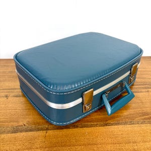 May include: A rectangular teal suitcase with a textured finish, featuring a matching teal handle, silver-toned metal latches, and white stitching along the edges. The suitcase has a silver-colored trim band running horizontally around the center.