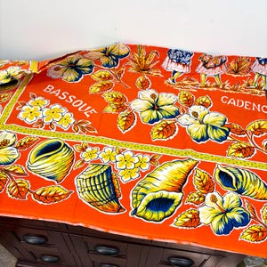 May include: An orange fabric with a floral and seashell pattern. The design includes yellow and white flowers, blue and yellow seashells, and the words "BASSOU" and "CADENCE". The fabric is folded over a dark wooden surface.