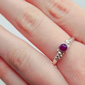 Puede incluir: Un anillo de plata con una peque&ntilde;a piedra preciosa redonda de color p&uacute;rpura engastada en el centro. El anillo est&aacute; adornado con peque&ntilde;as cuentas redondas en la banda.