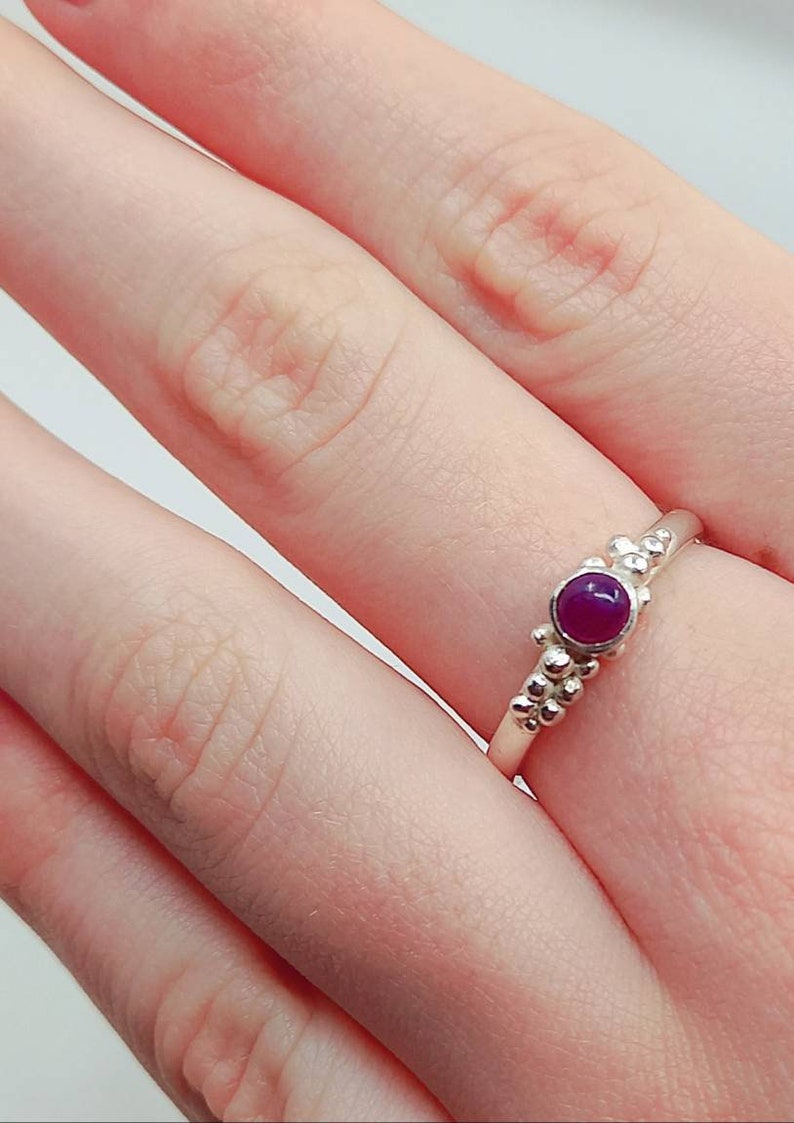Puede incluir: Un anillo de plata con una peque&ntilde;a piedra preciosa redonda de color p&uacute;rpura engastada en el centro. El anillo est&aacute; adornado con peque&ntilde;as cuentas redondas en la banda.