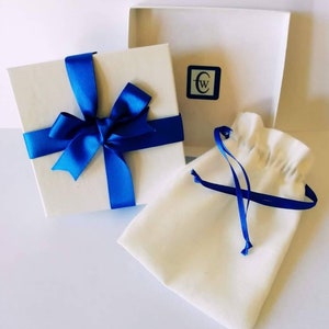 Puede incluir: Una caja de regalo blanca con una cinta y un lazo azules. La caja est&aacute; sobre una superficie blanca junto a una bolsa de tela blanca con una cinta azul.