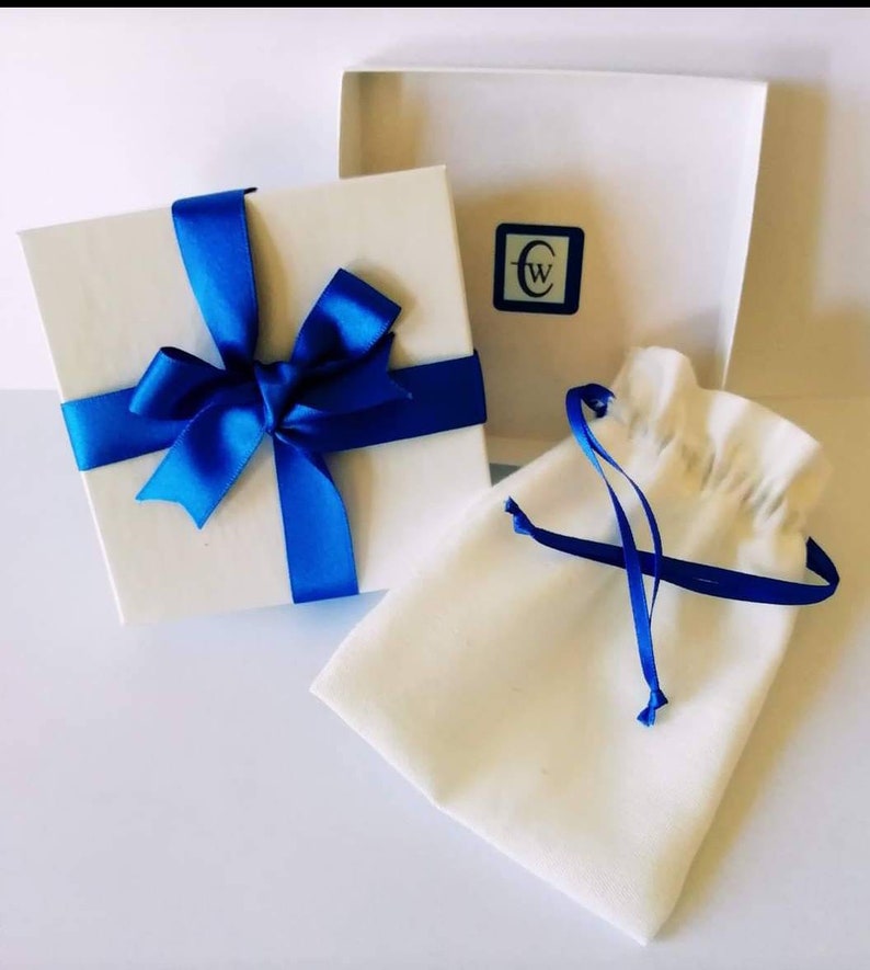 Puede incluir: Una caja de regalo blanca con una cinta y un lazo azules. La caja est&aacute; sobre una superficie blanca junto a una bolsa de tela blanca con una cinta azul.
