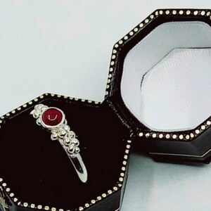 May include: A silver ring with a red gemstone set in a silver bezel. The ring is surrounded by small silver beads. The ring is in a black ring box with a white interior.
