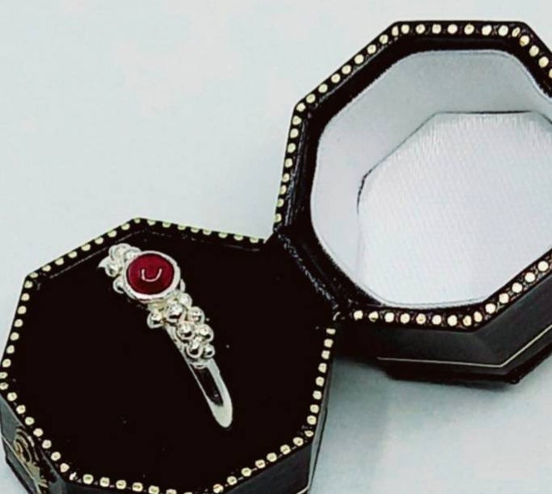 Puede incluir: Un anillo de plata con una piedra preciosa roja engastada en un engaste de plata. El anillo est&aacute; rodeado de peque&ntilde;as cuentas de plata. El anillo est&aacute; en una caja de anillo negra con un interior blanco.