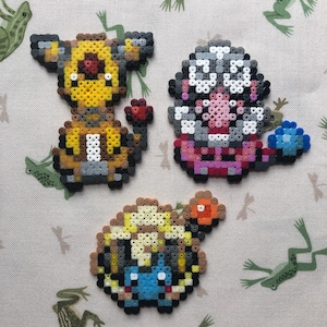 May include: Three colourful pixel art figures made from small plastic beads. The figures include a yellow and brown creature with a red nose, a pink and grey figure with a blue gem, and a blue and yellow creature. The figures are on a light green background.