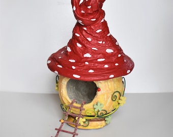 Op de afbeelding: Een fantasierijk feeënhuis gemaakt van papier-maché, gevormd als een paddenstoel met een rood-witte bovenkant en een gele onderkant. Het huis heeft een kleine deur en een ladder die er naartoe leidt. Er zijn groene wijnstokken en gele bloemen die het huis versieren.