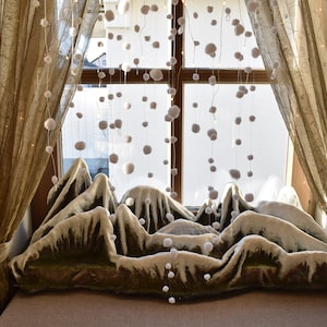 Może przedstawiać: Okno z przezroczystymi zasłonami i śnieżnym krajobrazem górskim wykonanym z tkaniny i pomponów wiszących z górnej części okna.