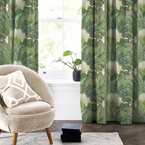 May include: A beige armchair with a white and beige patterned cushion sits in front of a window with green patterned curtains. The curtains feature a repeating pattern of large green leaves.