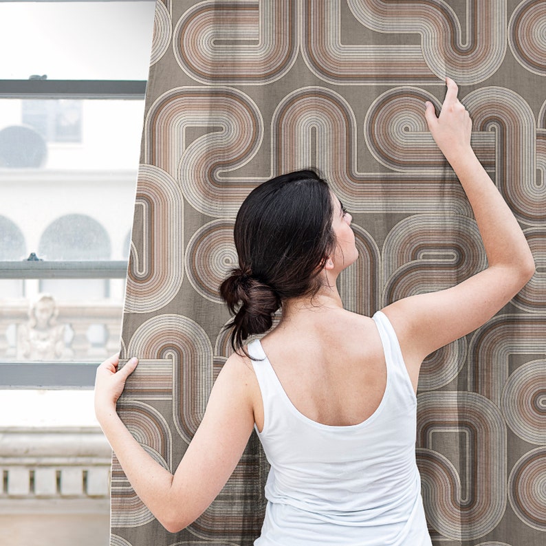 May include: A woman stands in front of a curtain with a geometric pattern of brown and beige curved lines. She is wearing a white tank top and has her back to the camera.