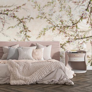 Op de afbeelding: Een slaapkamer met een bed met een roze hoofdbord, kussens en een dikke gebreide deken. De muur is versierd met bloemenbehang. Een nachtkastje met lades staat rechts. Het kleurenschema is zacht en gedempt.