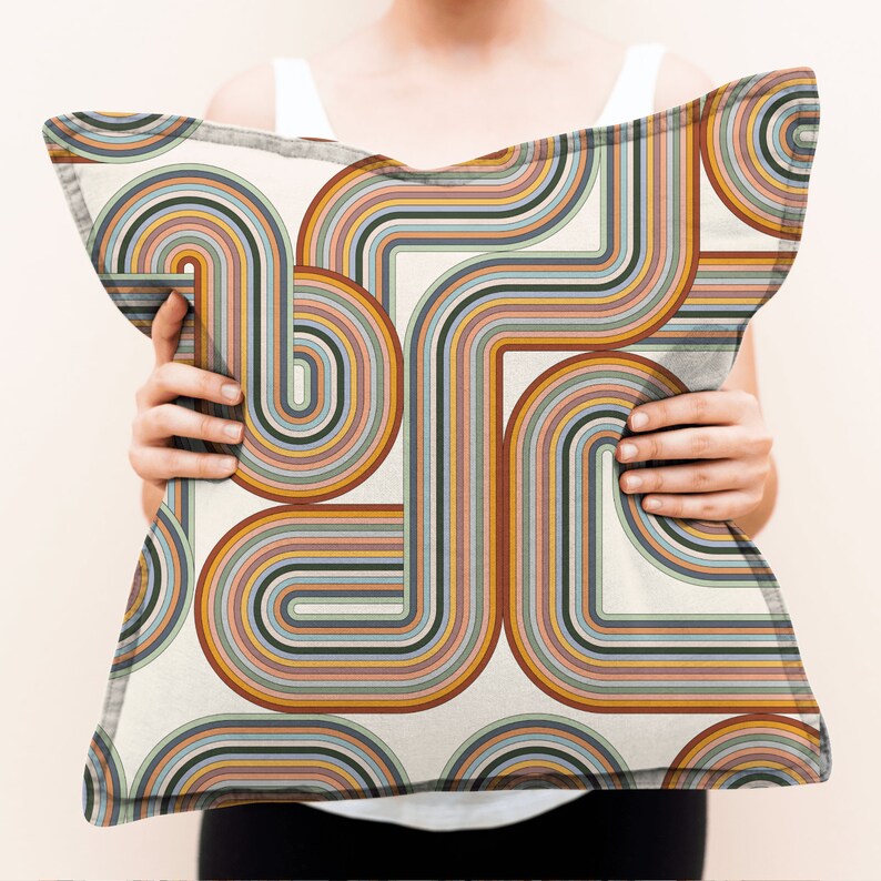 May include: A white cushion with a colourful geometric pattern of curved lines in shades of orange, yellow, blue, green, and brown. The pattern resembles a retro 70s design.