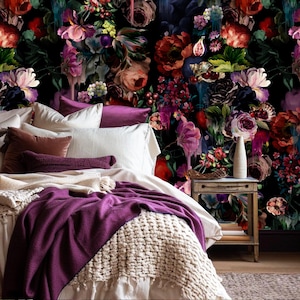 Może przedstawiać: Sypialnia z łóżkiem z białą pościelą, fioletowymi poduszkami i ciemnofioletowym kocem. Ściana zagłówka pokryta jest ciemną kwiatową tapetą z czerwonymi, różowymi i fioletowymi kwiatami. Mały drewniany stolik nocny trzyma wazon.