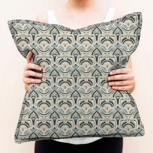 May include: Decorative pillow with a repeating geometric pattern in shades of teal, beige, and black. The pillow has a soft, textured appearance and is held up by a person.