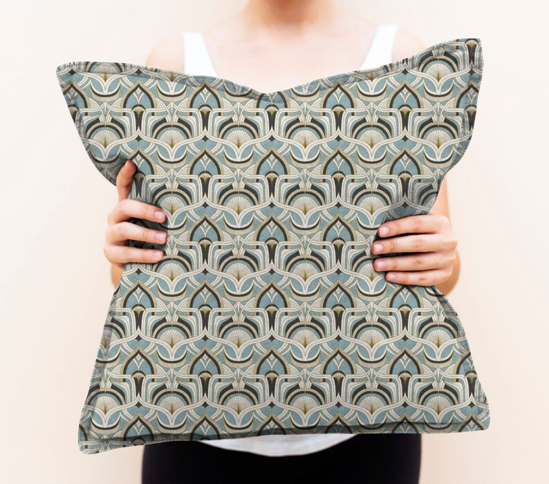 May include: Decorative pillow with a repeating geometric pattern in shades of teal, beige, and black. The pillow has a soft, textured appearance and is held up by a person.