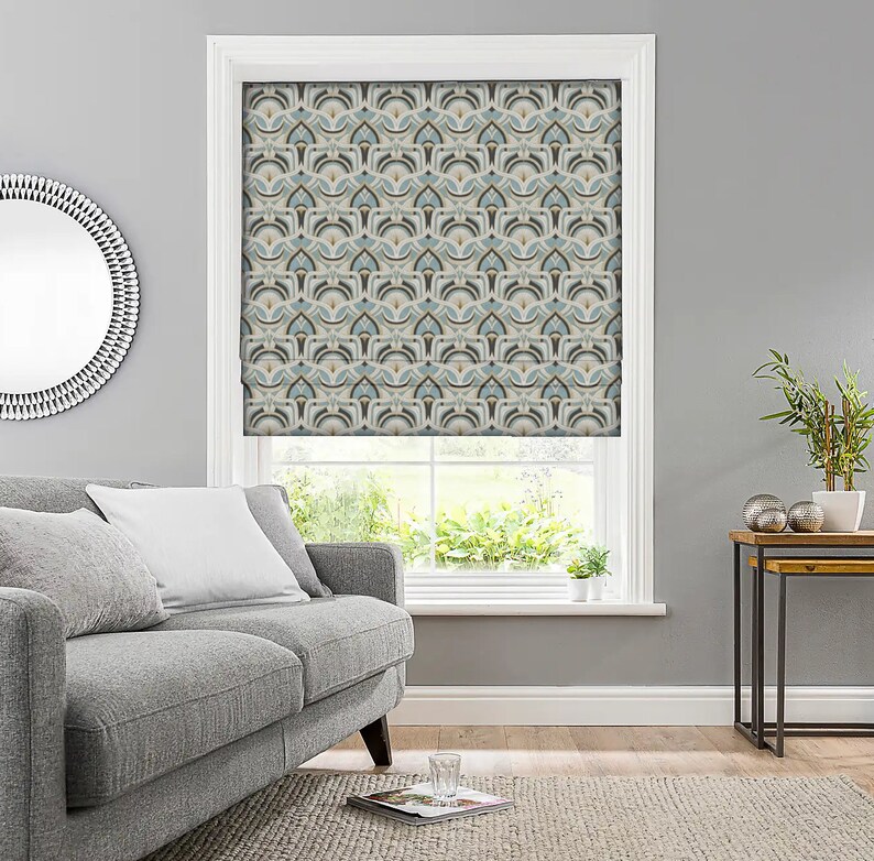 May include: A window with a patterned roller shade. The shade features a repeating geometric design in shades of blue, beige, and brown. The window is framed in white and the room has gray walls. A gray sofa and a round mirror are visible.