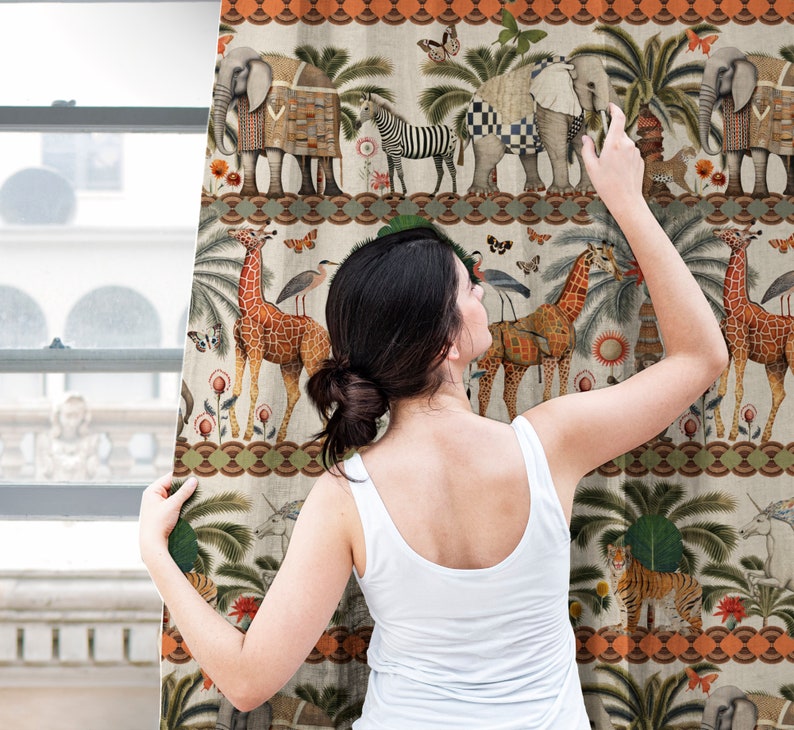 May include: A woman stands in front of a curtain featuring a colorful pattern of animals, plants, and geometric designs. The curtain has a cream background with orange, green, and blue accents. The animals include elephants, giraffes, zebras, and tigers.