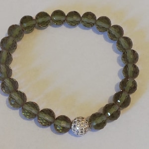 Könnte beinhalten: Ein grünes Kristallperlenarmband mit einem silbernen Kugelanhänger. Der Anhänger ist mit kleinen klaren Kristallen besetzt.