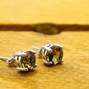May include: A pair of stud earrings with silver settings and round, faceted gemstones. The gemstones have a mix of olive green and golden brown hues. The earrings are displayed on a wooden surface.