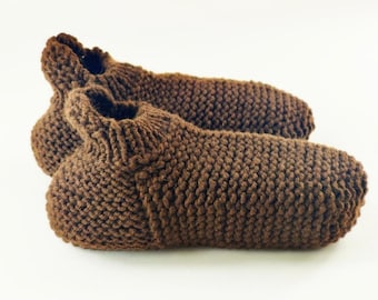May include: A pair of brown hand-knitted slippers featuring a textured garter stitch pattern throughout the body and a ribbed cuff around the ankle opening. The slippers are crafted from a thick yarn and have a rounded toe and heel design.