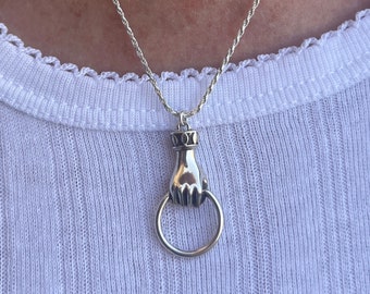 Könnte beinhalten: Eine silberfarbene Kordelkette mit einem Anhänger in Form einer Hand, die einen kreisförmigen Ring hält. Der Anhänger hat eine polierte metallische Oberfläche mit dunklen, vertieften Details am Handgelenk und hängt an einer dünnen Kette.