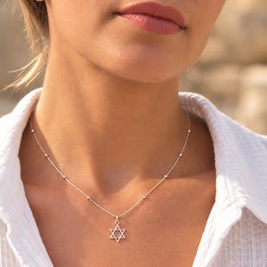 May include: A delicate silver necklace featuring a small Star of David pendant. The chain is composed of thin links interspersed with tiny, evenly spaced silver beads, creating a refined and subtle look that rests against the collarbone area.
