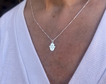 May include: A silver chain necklace with a hamsa hand charm. The charm has Hebrew writing on it.