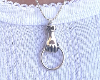Könnte beinhalten: Eine silberne Halskette mit einem handförmigen Anhänger und einem Ring. Die Hand hält den Ring.