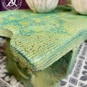 May include: A small, green wooden stool with a distressed finish. The stool has a unique, irregular shape and is painted in shades of green and blue. The stool is sitting on a patterned tile floor.