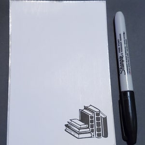 May include: A white notepad with a black and white illustration of a stack of books in the bottom right corner. A black Sharpie fine point permanent marker is on the right side of the notepad.
