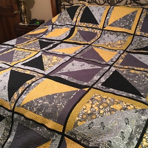 May include: A quilt featuring a geometric pattern in shades of grey, black, and yellow. The quilt is made from different fabrics with various patterns, including floral and paisley designs.