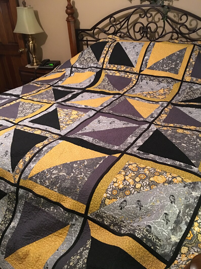 May include: A quilt featuring a geometric pattern in shades of grey, black, and yellow. The quilt is made from different fabrics with various patterns, including floral and paisley designs.
