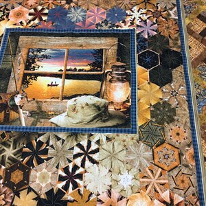 May include: A quilt with a blue and white checkered border and a central panel depicting a sunset scene through a window. The scene includes a lake, trees, and a boat with two figures. The quilt is made up of various geometric shapes in shades of brown, beige, and green.