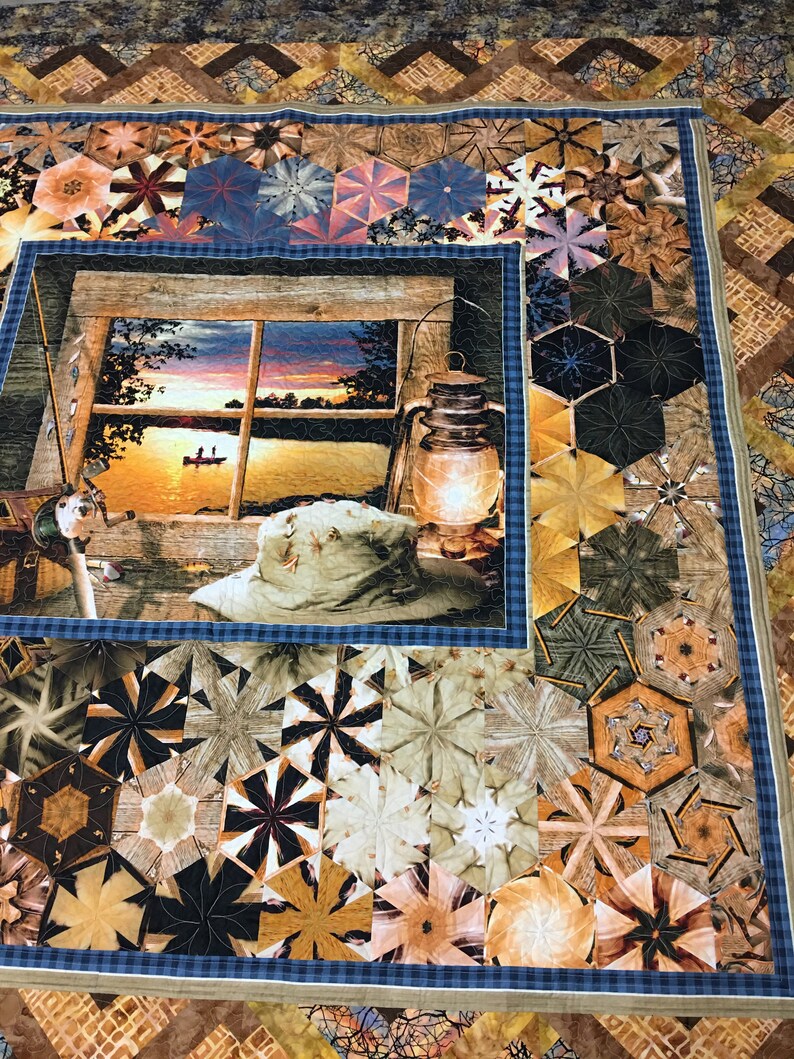 May include: A quilt with a blue and white checkered border and a central panel depicting a sunset scene through a window. The scene includes a lake, trees, and a boat with two figures. The quilt is made up of various geometric shapes in shades of brown, beige, and green.