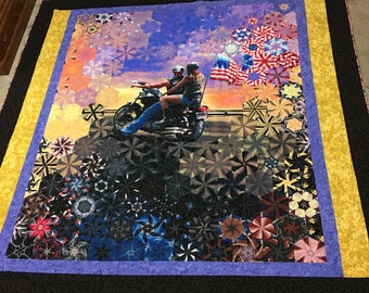 May include: A quilt featuring a couple riding a motorcycle with an American flag waving in the background. The quilt is made of various fabrics with a geometric pattern and a black border.