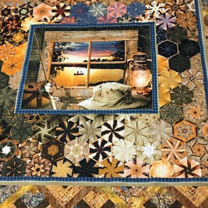 May include: A quilt with a patchwork design featuring a variety of geometric shapes in shades of brown, tan, and blue. The center of the quilt features a framed image of a lake scene with a sunset, a boat, and a lantern.