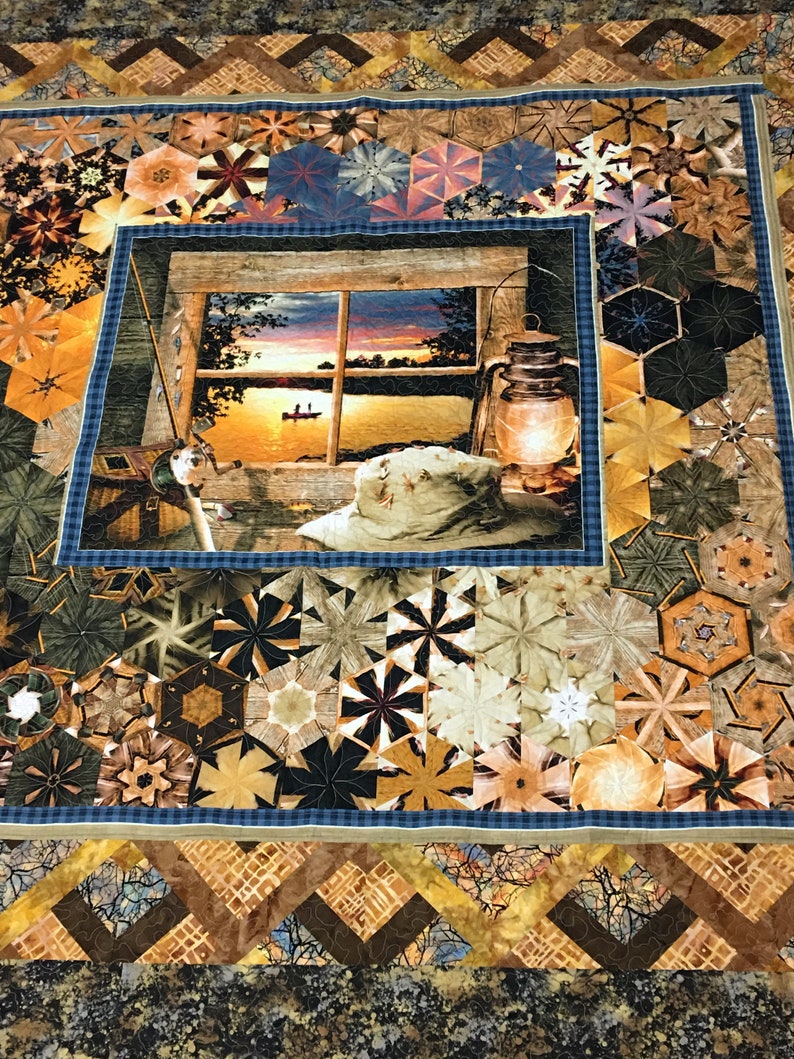 May include: A quilt with a patchwork design featuring a variety of geometric shapes in shades of brown, tan, and blue. The center of the quilt features a framed image of a lake scene with a sunset, a boat, and a lantern.