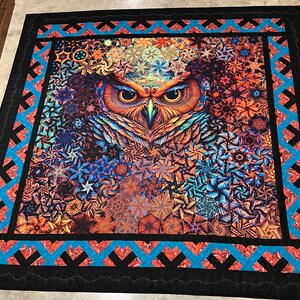 May include: A colorful quilt with a large owl design in the center. The owl has a detailed pattern of feathers and a bright orange beak. The quilt is surrounded by a black border with a blue and red geometric pattern.