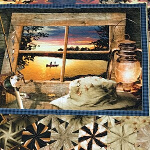 May include: A quilt with a patchwork design featuring a variety of geometric shapes in shades of brown, tan, and blue. The center panel depicts a window with a view of a lake at sunset, with two figures in a boat on the water. A lantern and a hat are also visible in the scene.