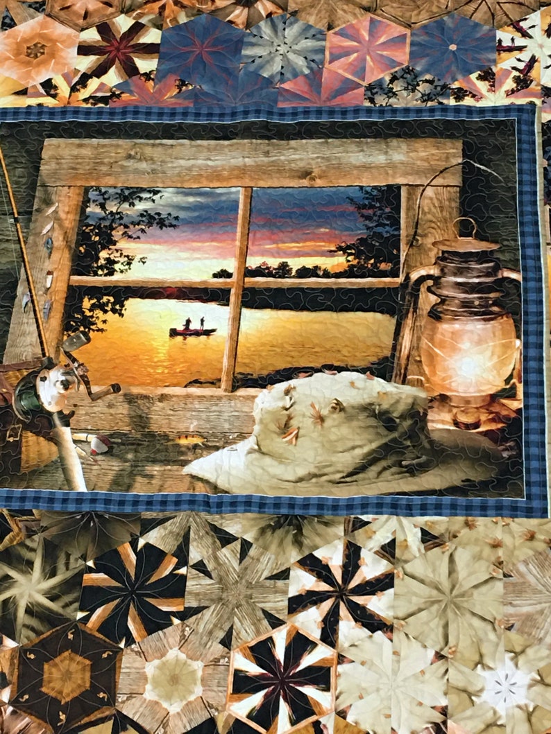 May include: A quilt with a patchwork design featuring a variety of geometric shapes in shades of brown, tan, and blue. The center panel depicts a window with a view of a lake at sunset, with two figures in a boat on the water. A lantern and a hat are also visible in the scene.